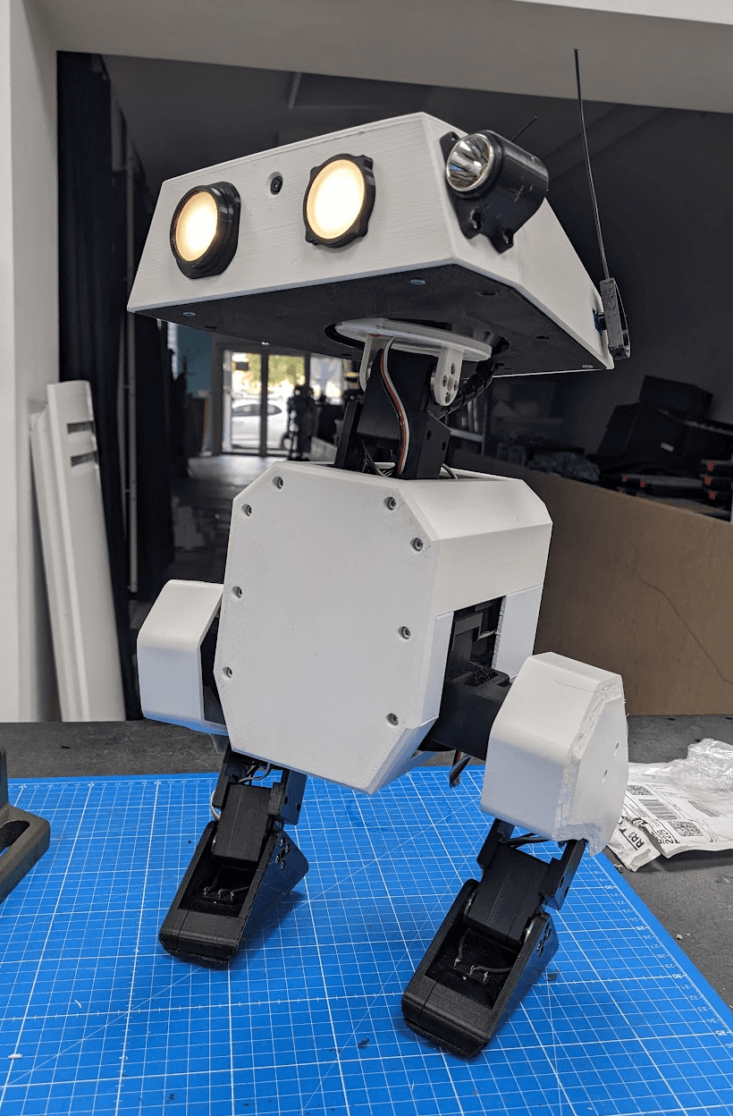 Open Duck Mini v2: white 3D-printed body and head with two lit eyes, on black legs, standing on a cutting mat
