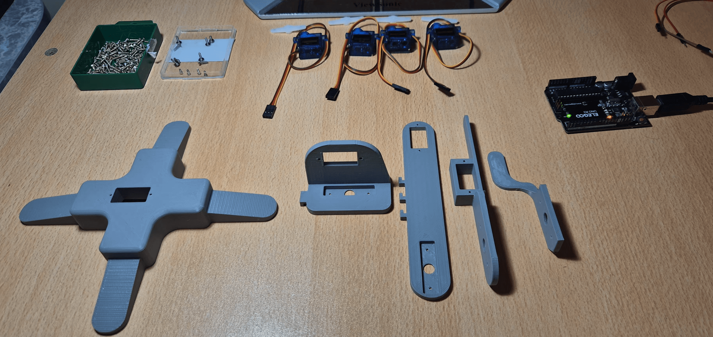 Printed arm components, servos, screws and an Arduino board laid out on a table.