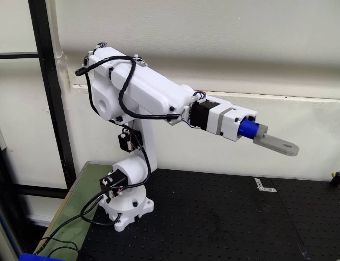 A white robotic arm mounted on a workbench, shown with its cables and end attachment.