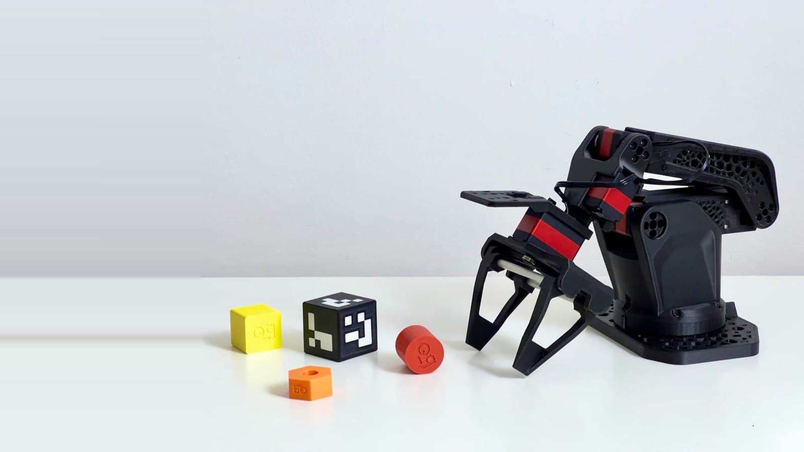 A black robot arm with a parallel gripper beside colored objects on a white tabletop.