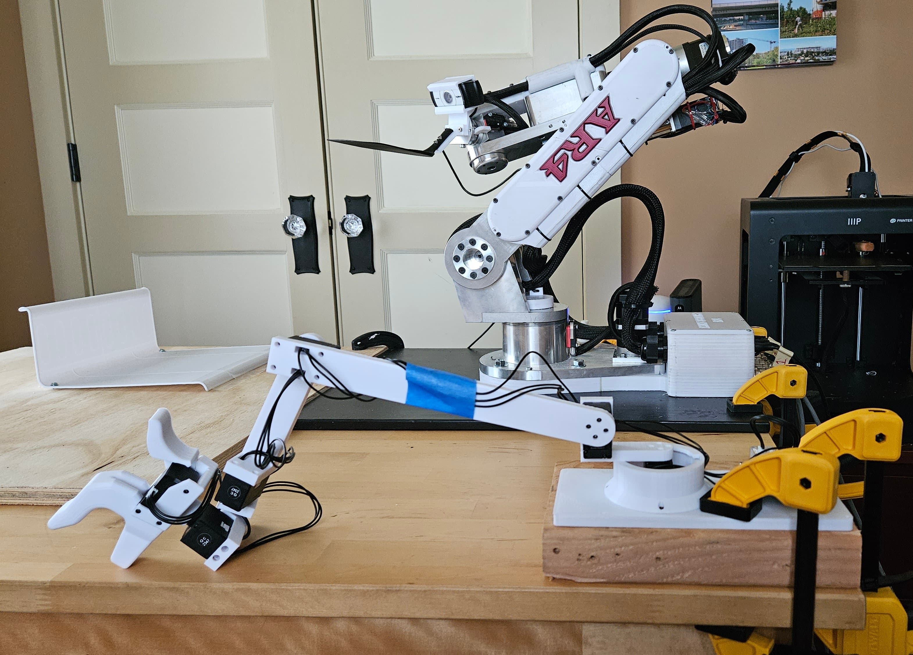 A white articulated GELLO arm with a gripper in front of a larger articulated robot arm on a table.